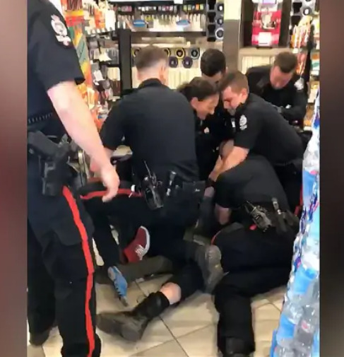 A screengrab from the bystander video of the incident, showing six police officers pinning a person to the floor, with another officer standing in the foreground