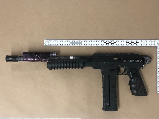 A photo of a weapon on a brown background alongside a ruler. Something is taped on the barrel, possibly a flashlight.