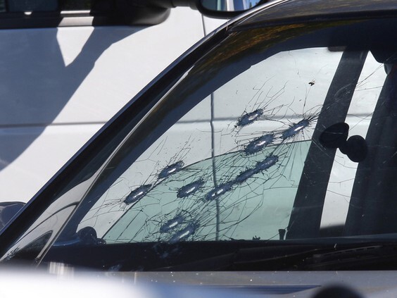 A photo of what appears to be the passenger side of the front windshield of a white vehicle; 11 bullet holes are visible in the windshield.