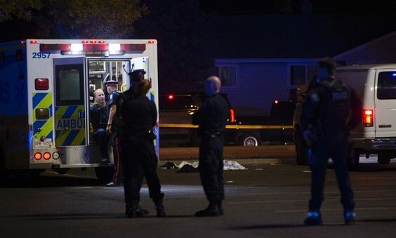 A photo of the crime scene at night. A body covered by a white tarp lies on the ground between a white van and an ambulance. First responders are gathered nearby and in the back of the ambulance.