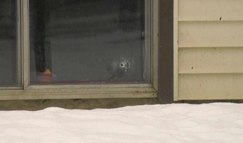 A close-up of a basement apartment window with a bullet hole through the glass. A superman figurine sits inside the windowsill.